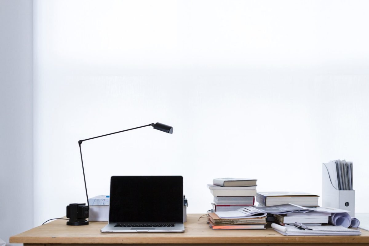 Minimalist desk with desk lamp, Apple Macbook, stack of books and papers against a white wall