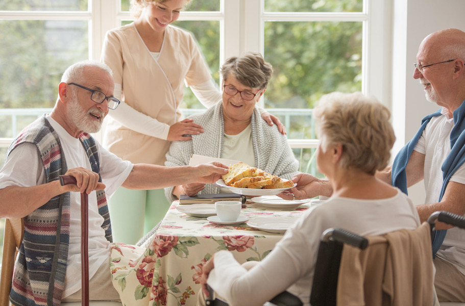 Group of four senior citizens eating a meal while a nurse stands beside their table