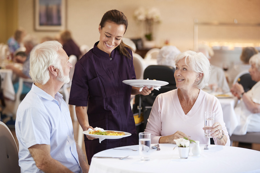 Senior couple dining in a long-term care facility, they are being served by a staff member in a purple dress.